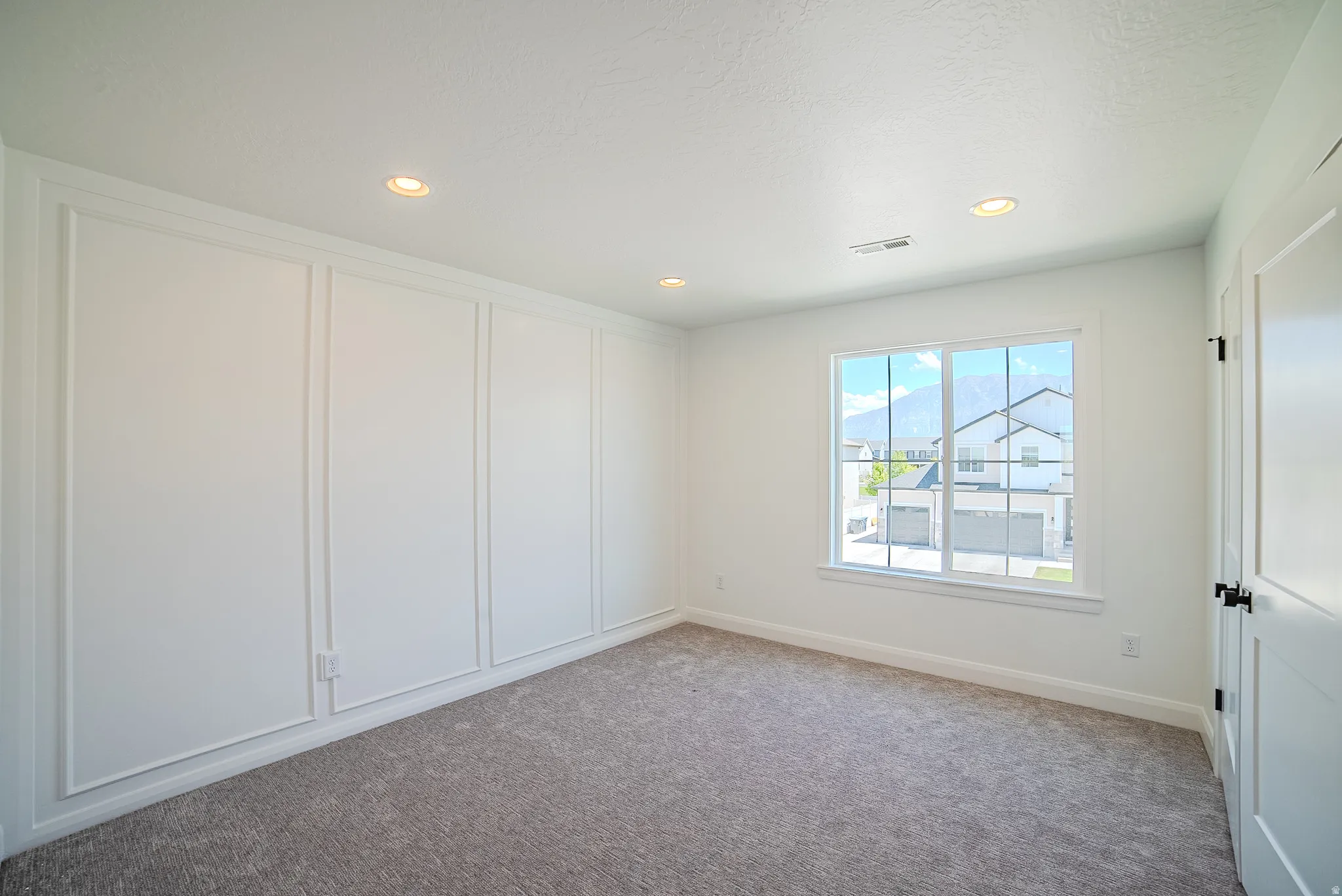 Carpeted interior room featuring a window with grid panes, recessed ceiling lighting, a paneled accent wall, and a white interior door with dark hardware