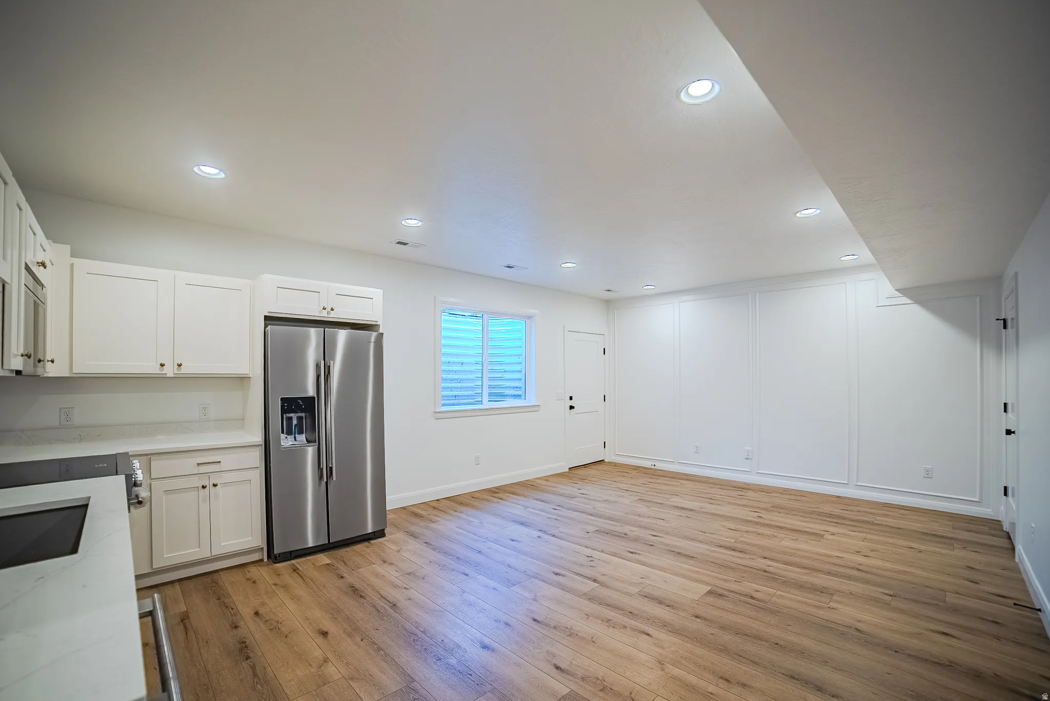 Open-concept living space featuring wood-finish flooring, recessed lighting, and a kitchen area with white cabinetry, a stainless steel refrigerator, and light-toned countertops