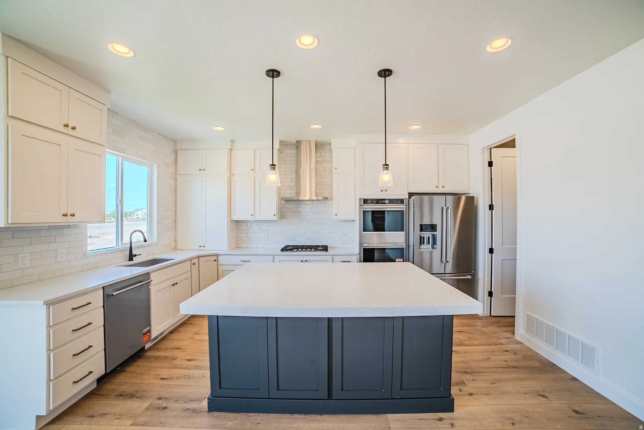 Contemporary kitchen featuring a large central island with a light-toned countertop, white cabinetry with gold-toned hardware, stainless steel appliances, wood-finish flooring, and recessed lighting