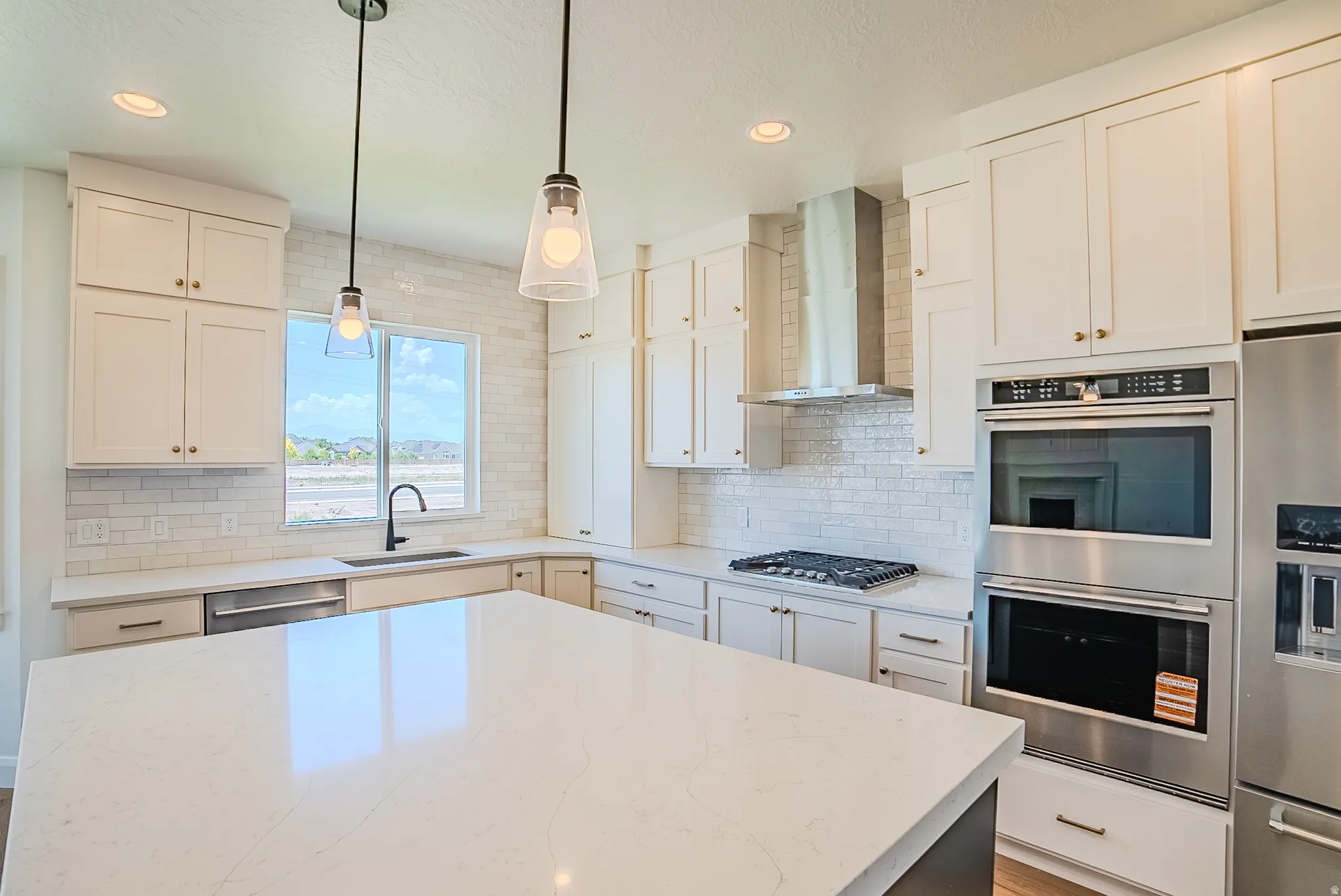 Modern kitchen featuring a spacious island with a light-toned countertop, white cabinetry with gold-toned hardware, stainless steel appliances including a built-in double oven and refrigerator, and a white subway tile backsplash