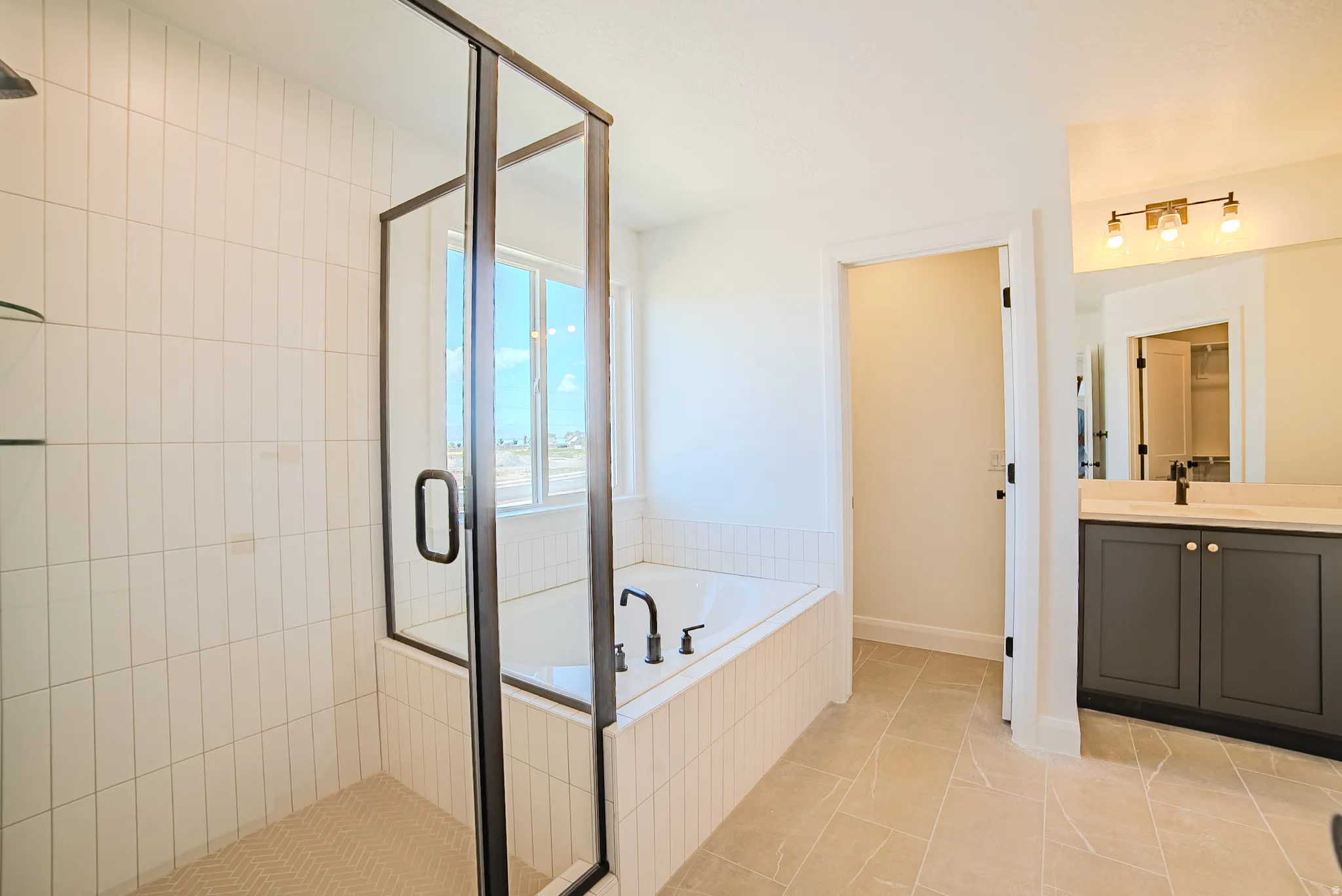 Contemporary bathroom featuring a walk-in shower with vertical subway tile, a built-in soaking tub, a dark gray vanity with a light-colored countertop, and a large window providing ample natural light