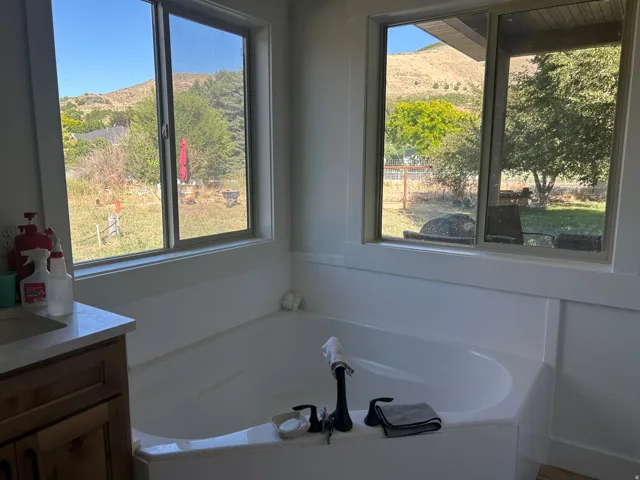Corner soaking tub with matte black fixtures, quartz vanity countertop, wood-finish cabinetry, and expansive window views of surrounding hills and mature trees