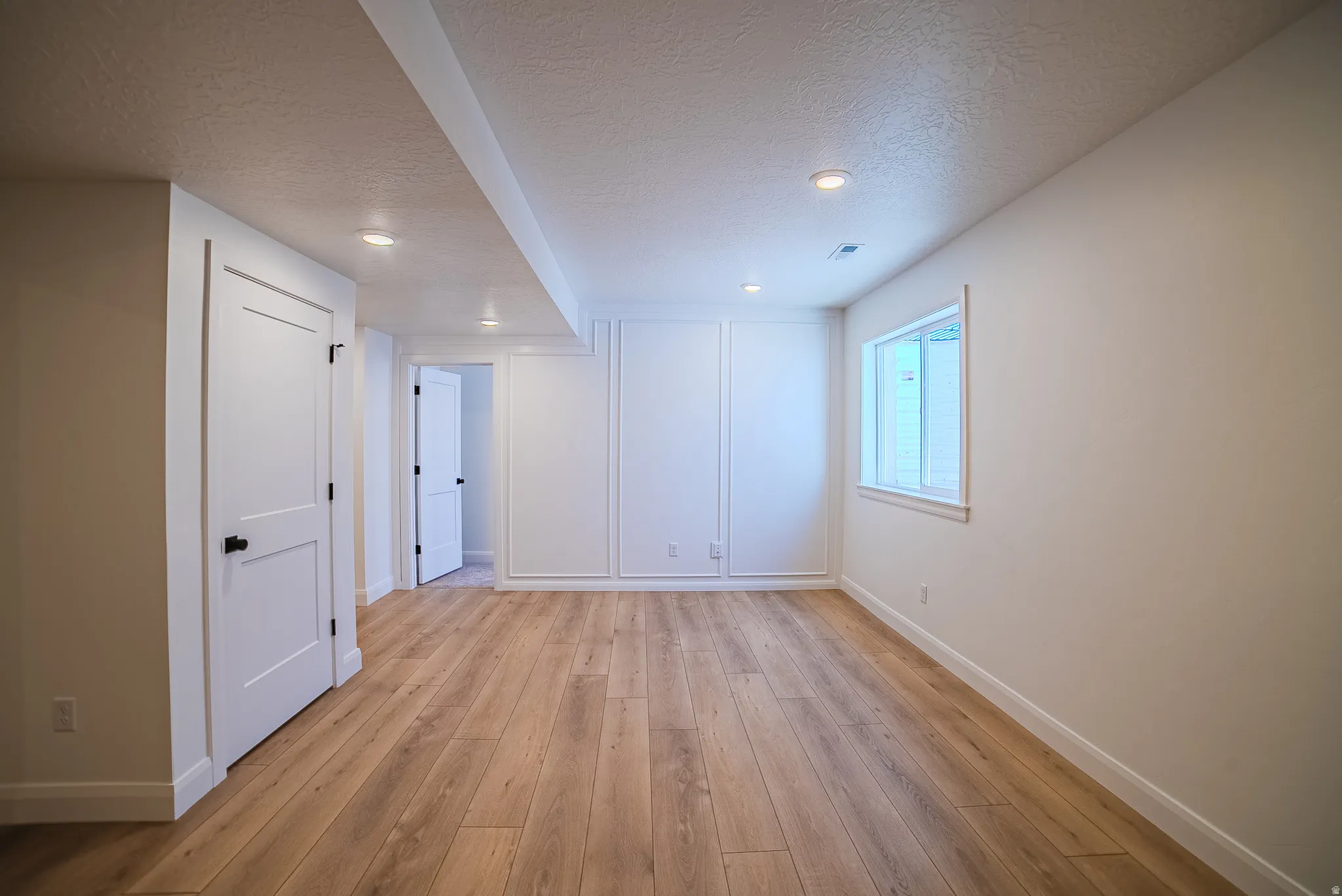 Spacious room featuring wood-finish flooring, recessed lighting, white doors with matte black hardware, and an egress window
