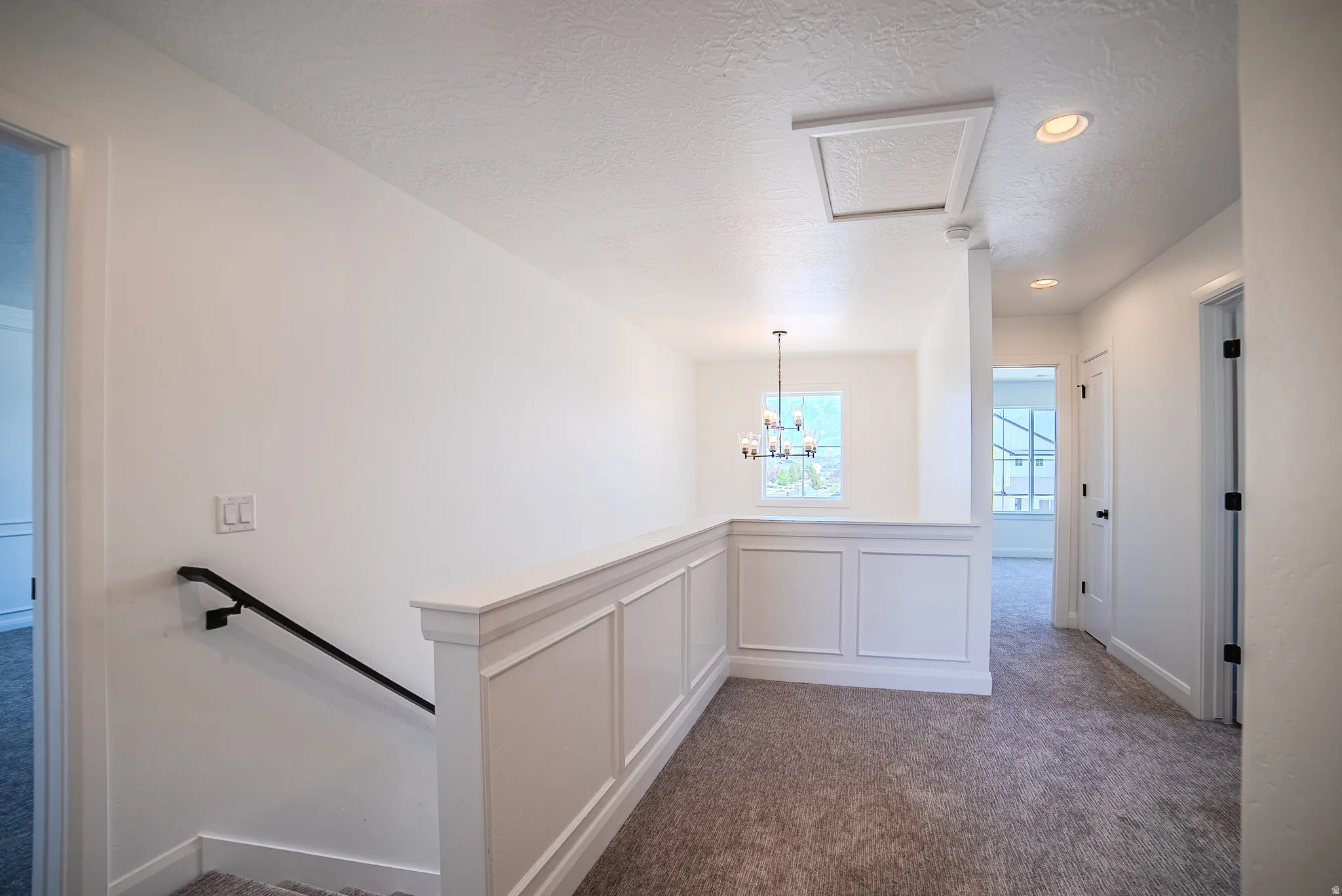 Upper-level landing featuring a white wainscoting half-wall, neutral carpeting, recessed ceiling lighting, and a modern chandelier visible through an opening