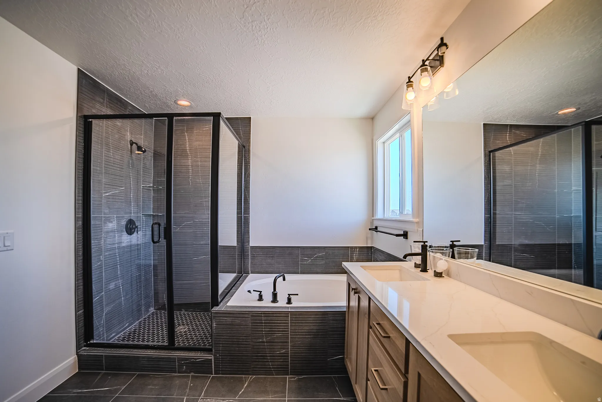 Modern bathroom featuring a double vanity with stone-finish countertops, an integrated soaking tub, a glass-enclosed shower with dark tile surround, and dark-finish fixtures