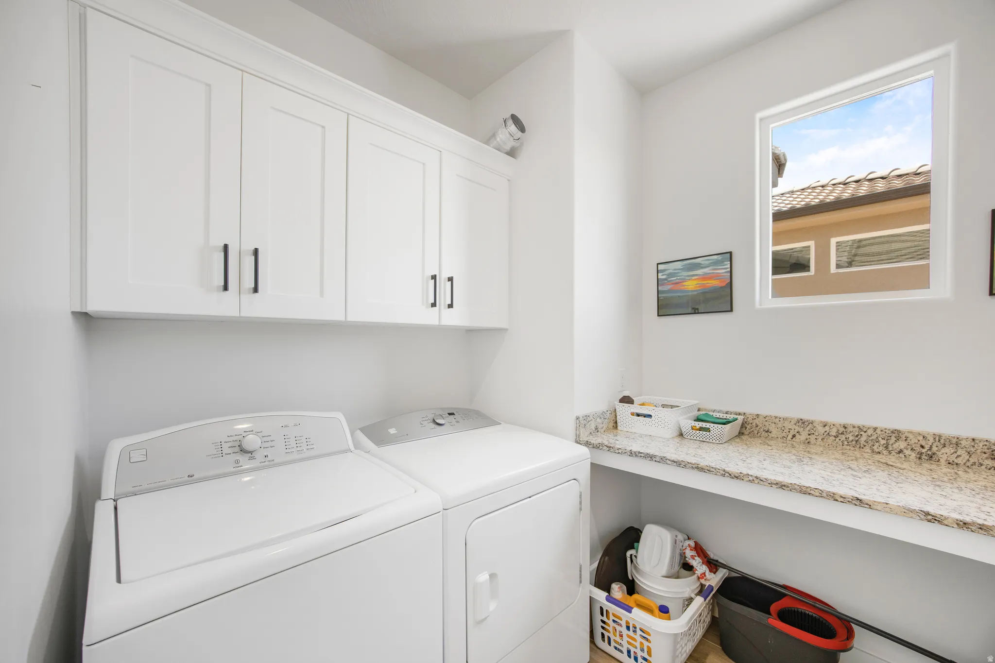 Dedicated laundry space featuring white shaker-style cabinetry with dark hardware, a spacious light-toned speckled countertop, an integrated window, and light-toned flooring