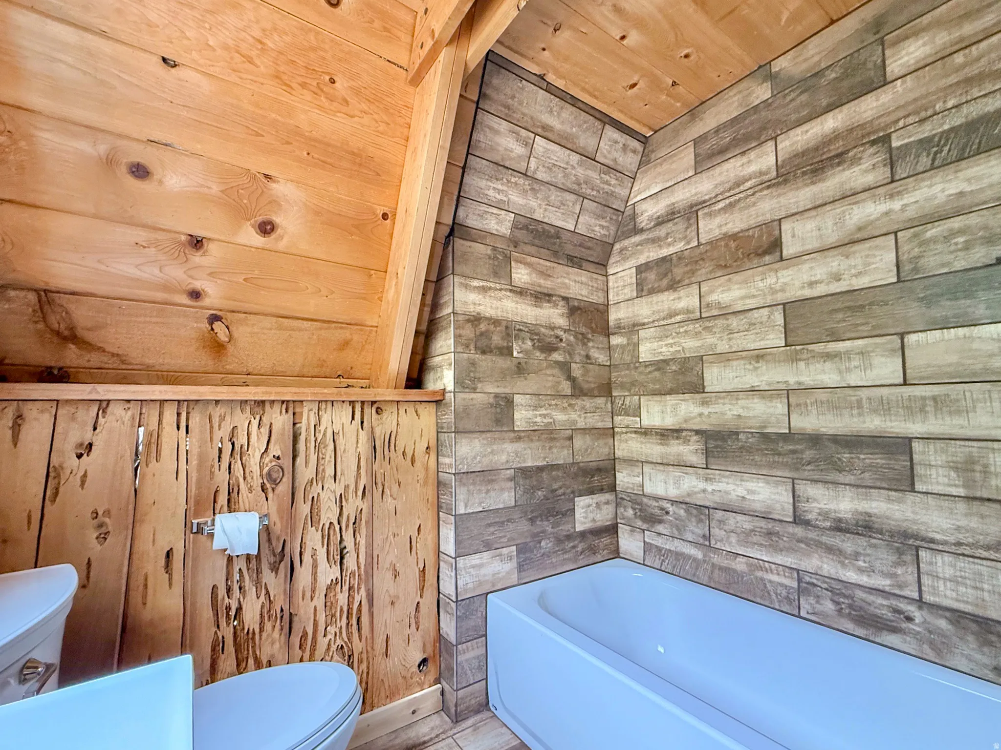 Compact bathroom featuring a white bathtub, wood-finish tile shower surround, tongue-and-groove wood paneling, and a unique wood plank accent wall