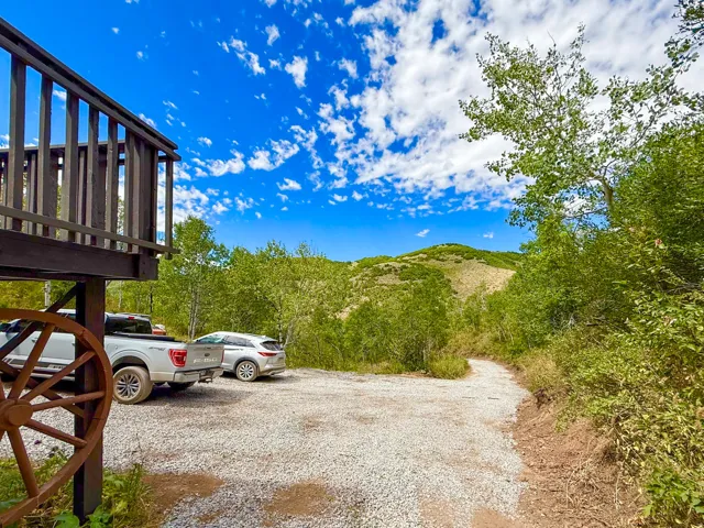 Elevated wood deck with baluster railing, gravel parking area, and a winding dirt path surrounded by mature trees and natural landscaping