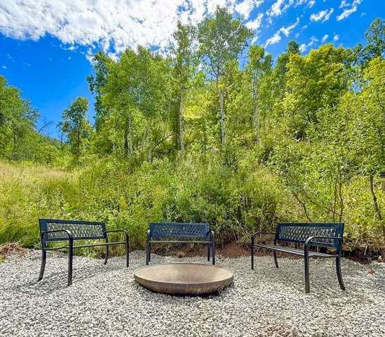Gravel outdoor seating area featuring a metal fire pit and three metal benches, surrounded by mature trees and natural landscaping
