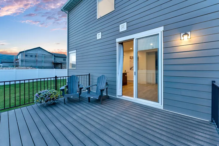 Spacious exterior deck with composite decking and black metal railings, featuring sliding glass door access, exterior wall lighting, and a fenced yard