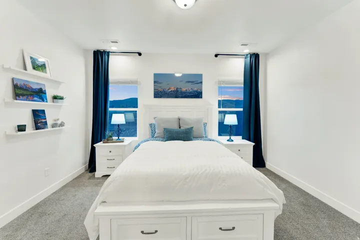 Bright bedroom featuring two windows with dark blue drapery, a white paneled headboard, built-in shelving, and light gray carpeting