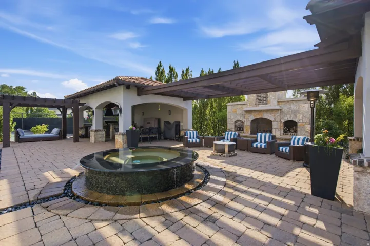 Expansive paver patio with a central water feature, covered outdoor kitchen, stone fireplace, and a large pergola