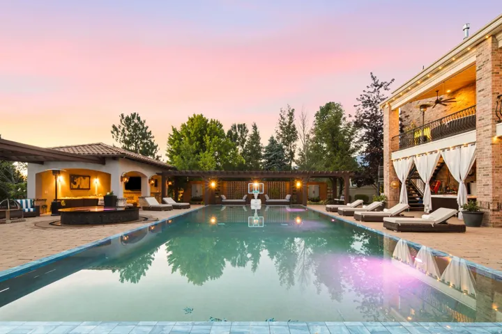 Expansive outdoor space featuring a rectangular swimming pool, a covered patio structure with a tile roof, an integrated fire pit, and a multi-level brick exterior