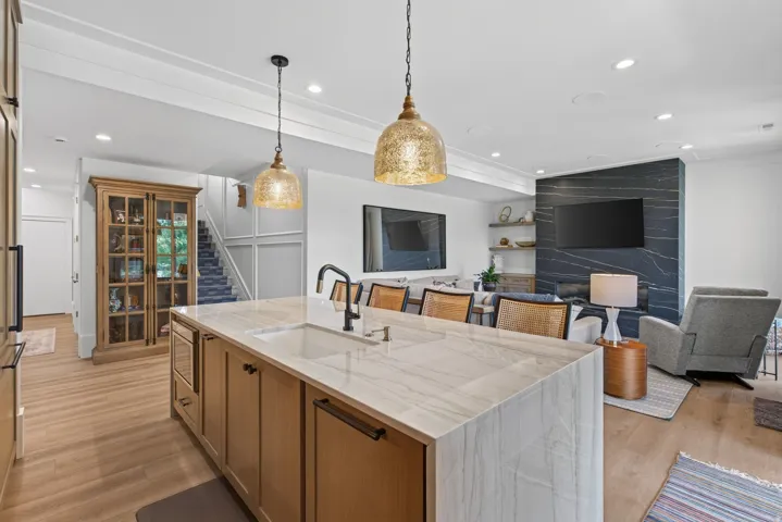Guest House Open-concept living area featuring a kitchen island with integrated sink, wood-finish flooring, a stone-clad fireplace surround, recessed lighting, and decorative pendant light fixtures