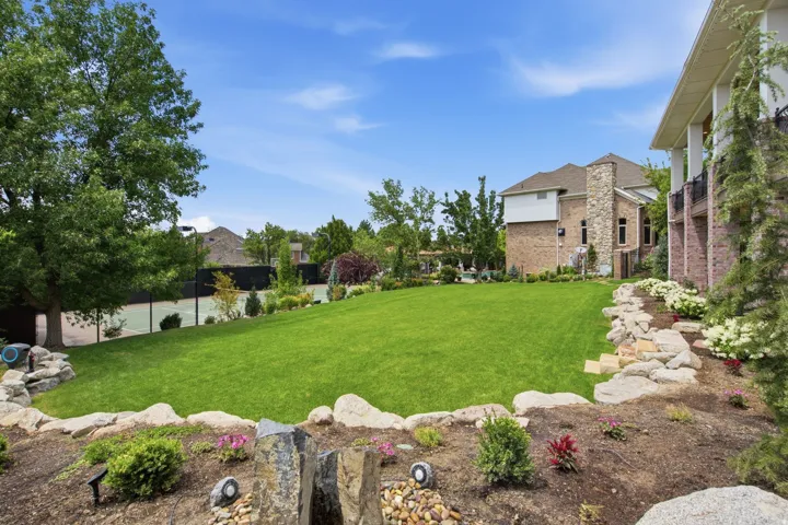 Guest House Expansive terraced lawn featuring mature landscaping, a private tennis court, and a stone-accented exterior with a covered balcony