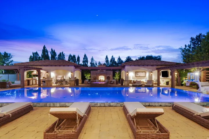 Expansive outdoor living area featuring a rectangular swimming pool with integrated lighting, two covered cabanas with arched entryways, and a central outdoor fireplace structure