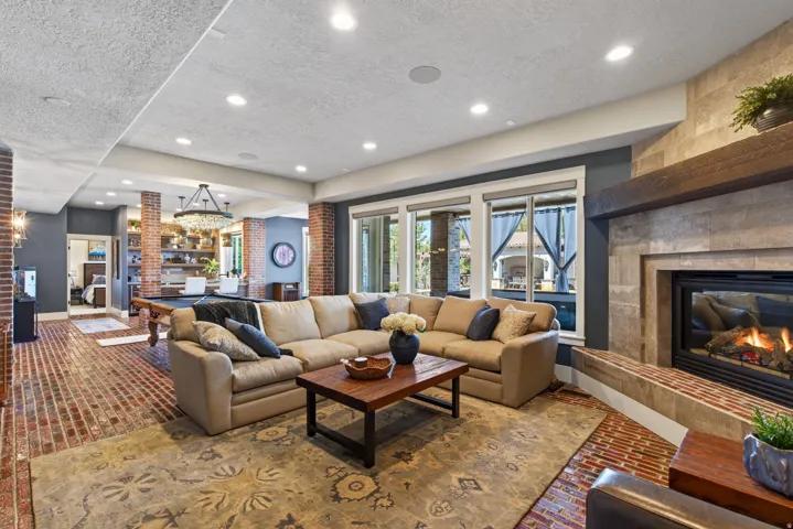 Spacious living area featuring a large stone-clad fireplace with a substantial wood mantel, recessed lighting, brick flooring, and multiple windows