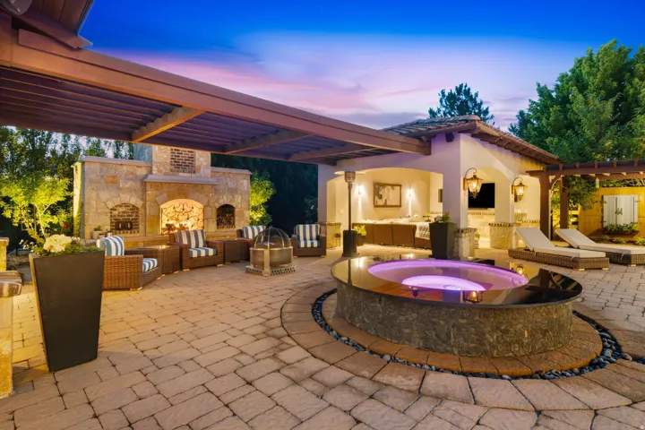Expansive paver patio featuring a built-in spa with stone surround, a stone outdoor fireplace, and a covered outdoor kitchen area with built-in cabinetry