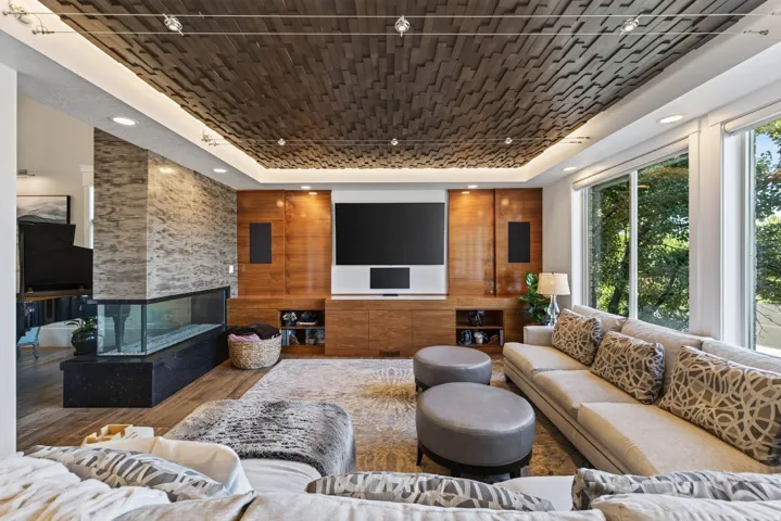 Living area featuring a textured wood-plank ceiling, large picture windows, a linear fireplace with a stone-clad surround, wood-finish flooring, and a built-in media center with wood paneling