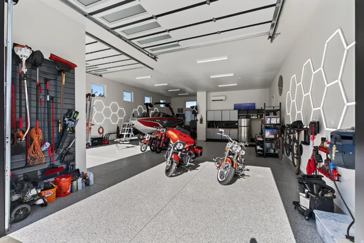 Spacious garage featuring a durable epoxy-coated floor, multiple storage systems, wall-mounted hexagonal lighting, and high ceilings with natural light panels