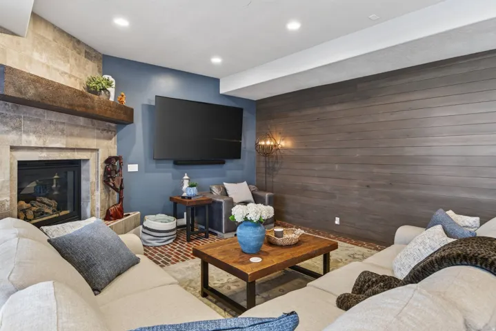 Living area featuring a stone-clad fireplace with a substantial wood mantel, recessed lighting, a wide plank wood-finish accent wall, and perimeter brick flooring
