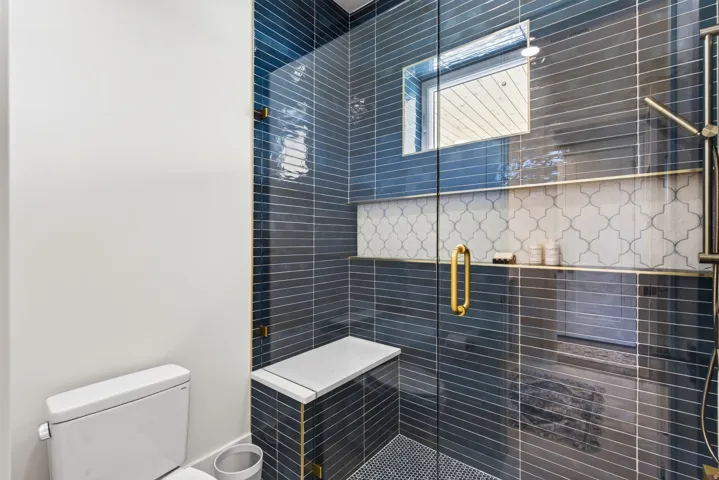 Guest House Custom tiled shower enclosure featuring dark blue horizontal subway tiles, a mosaic tile accent band, a built-in bench, and a gold-toned shower fixture