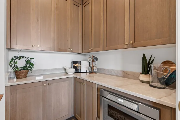 Guest House Kitchenette or pantry area featuring light wood-finish cabinetry with brass hardware, light-toned stone countertops, and a built-in stainless steel microwave