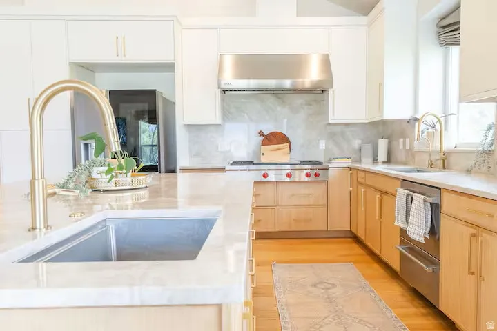 Light-toned kitchen featuring wood-finish flooring, light wood cabinetry, white upper cabinetry, stainless steel appliances, and a light-toned countertop with an undermount sink and a gold-toned faucet