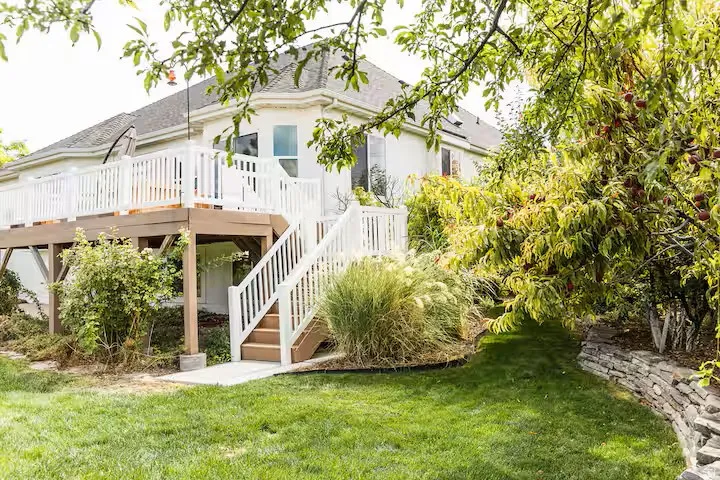 Expansive backyard featuring a multi-level deck with white railings, a stepped concrete walkway, lush green lawn, mature trees, and a stone retaining wall