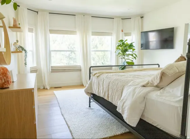 Room featuring wood-finish flooring, multiple windows with white drapery, and a wall-mounted television