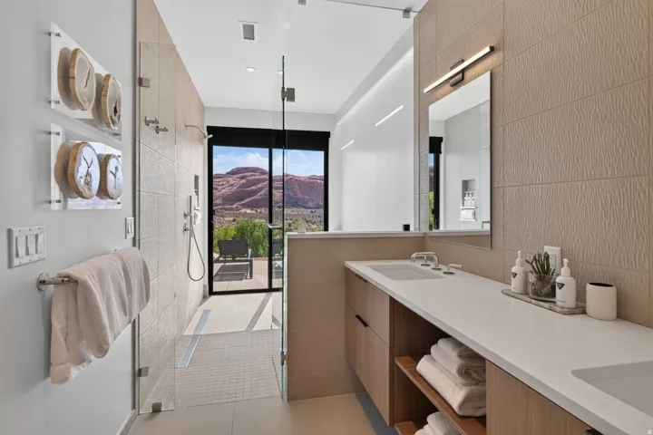 Contemporary bathroom featuring a dual vanity with light-tone wood-finish cabinetry, a large walk-in shower with glass enclosure, textured wall tiling, and a floor-to-ceiling window offering scenic natural rock formations
