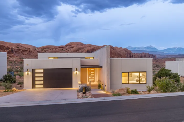 Modern architectural exterior with a flat roofline, integrated garage with frosted glass panels, illuminated entryway, and desert landscaping