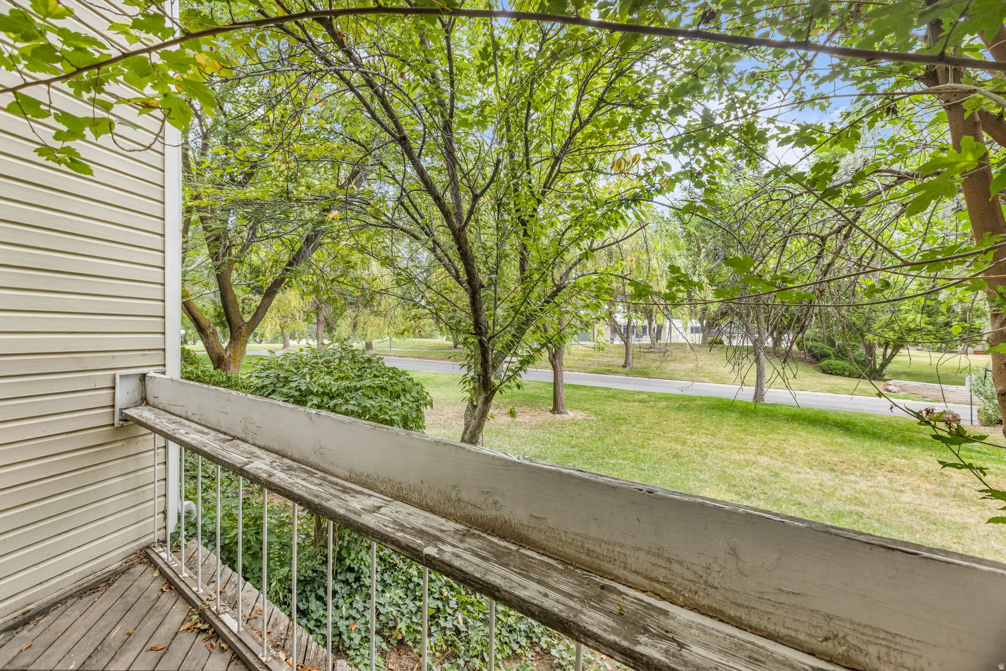 Private balcony with wood-finish decking and white metal railings, offering verdant tree-lined surroundings and a paved pathway