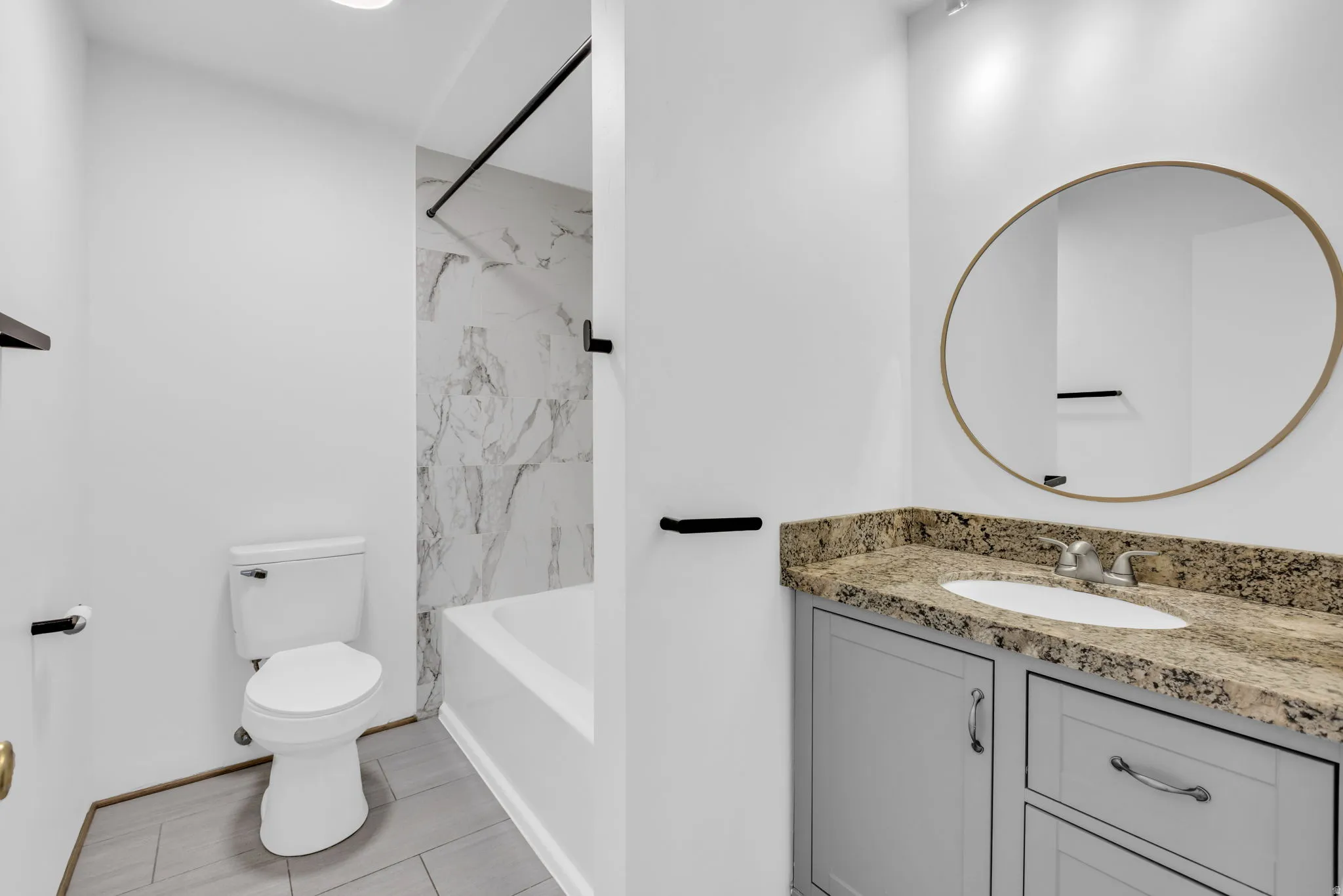 Bathroom featuring a light gray vanity with a granite countertop, oval-framed mirror, and a tub-shower combination with marble-look tile surround