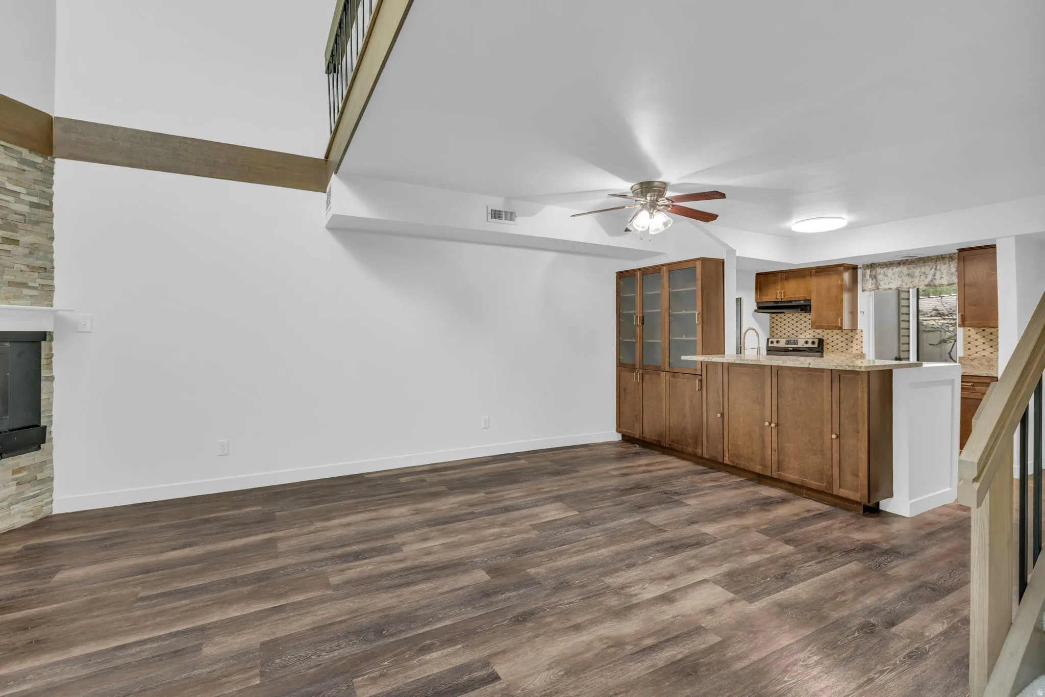 Open-concept living area with wood-finish flooring, a stacked stone fireplace, a kitchen island with stone countertop, wood cabinetry, and a ceiling fan