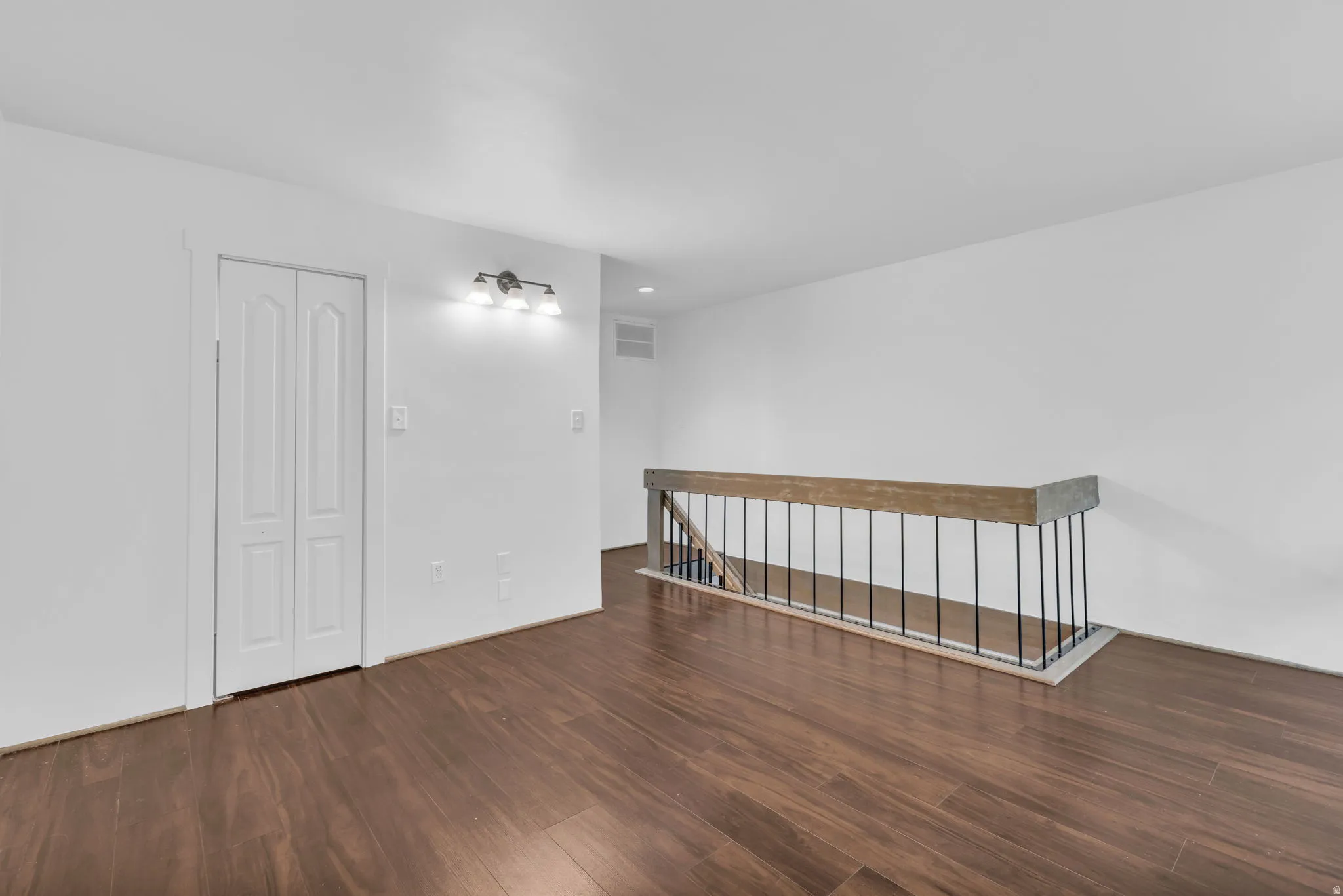 Open-concept area featuring rich wood-finish flooring, white walls, a contemporary three-light wall sconce, and a wood railing with black metal spindles
