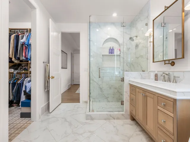 Bathroom featuring a glass-enclosed shower with a recessed niche, a single vanity with a light wood finish, a rectangular mirror with a brass-finish frame, and marble-look tile flooring