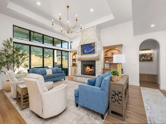 Spacious living area featuring a stone-clad fireplace, high ceilings with a tray detail, expansive floor-to-ceiling windows, wood-finish flooring, and a built-in bookshelf