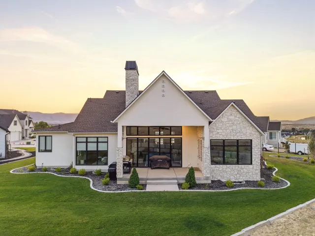 Contemporary exterior showcasing a covered patio with recessed lighting, expansive black-framed windows, a stone chimney, and a meticulously landscaped lawn with decorative rock beds