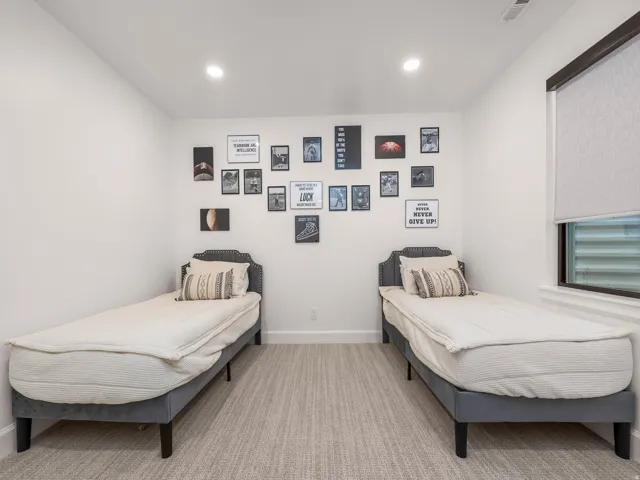 Recessed lighting illuminates this room featuring two upholstered headboards, a window with a roller shade, and textured carpeting
