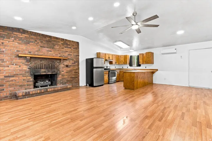 Open-concept living area with wood-finish flooring, a brick fireplace, and a kitchen featuring wood cabinetry, stainless steel appliances, and a breakfast bar