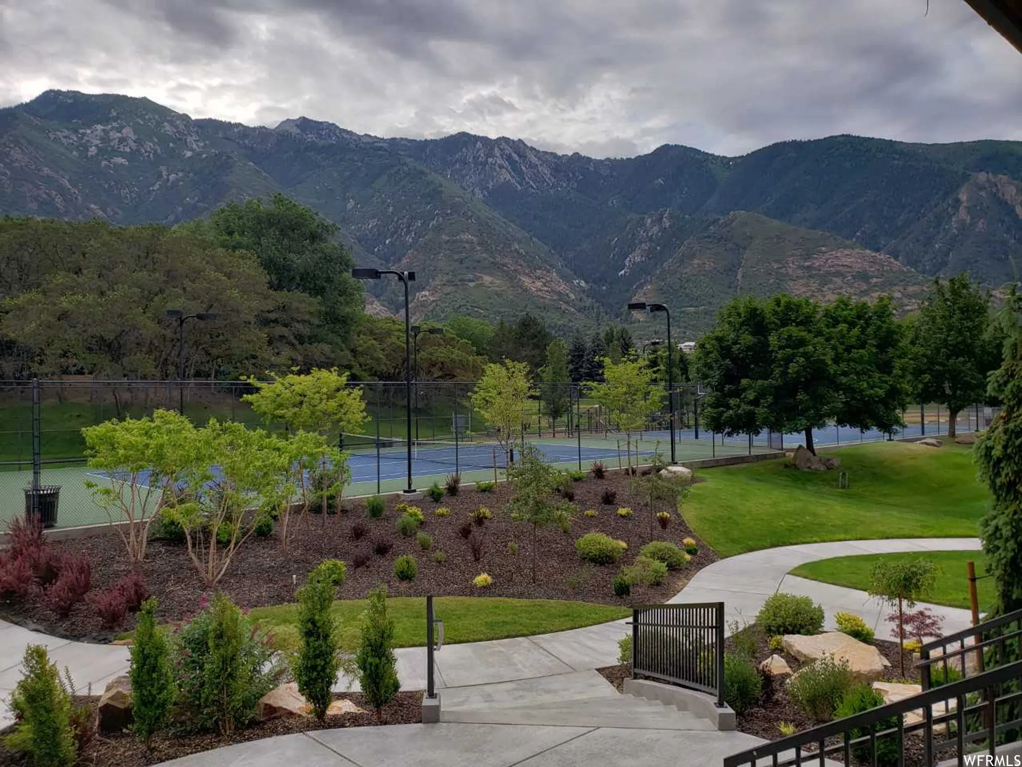 Professional tennis courts set against a mountain backdrop, featuring landscaped gardens, manicured lawns, and concrete pathways with modern railings