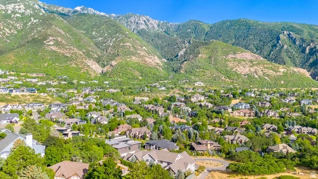 Residential neighborhood nestled at the base of verdant mountains, featuring various architectural styles, ample mature trees, and a clear blue sky