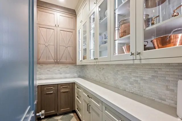 Spacious pantry featuring extensive built-in cabinetry, glass-front upper cabinets with interior lighting, light-toned stone countertops, and a full-height tile backsplash