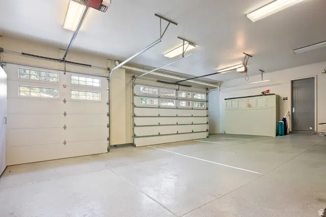 Three-car garage featuring white sectional doors with upper-panel windows, exposed overhead garage door openers, painted concrete flooring, and built-in white cabinetry