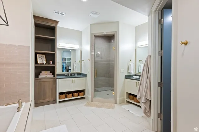 Spacious bathroom featuring a glass-enclosed shower, dual vanity sinks with dark countertops, built-in shelving, and light tile flooring