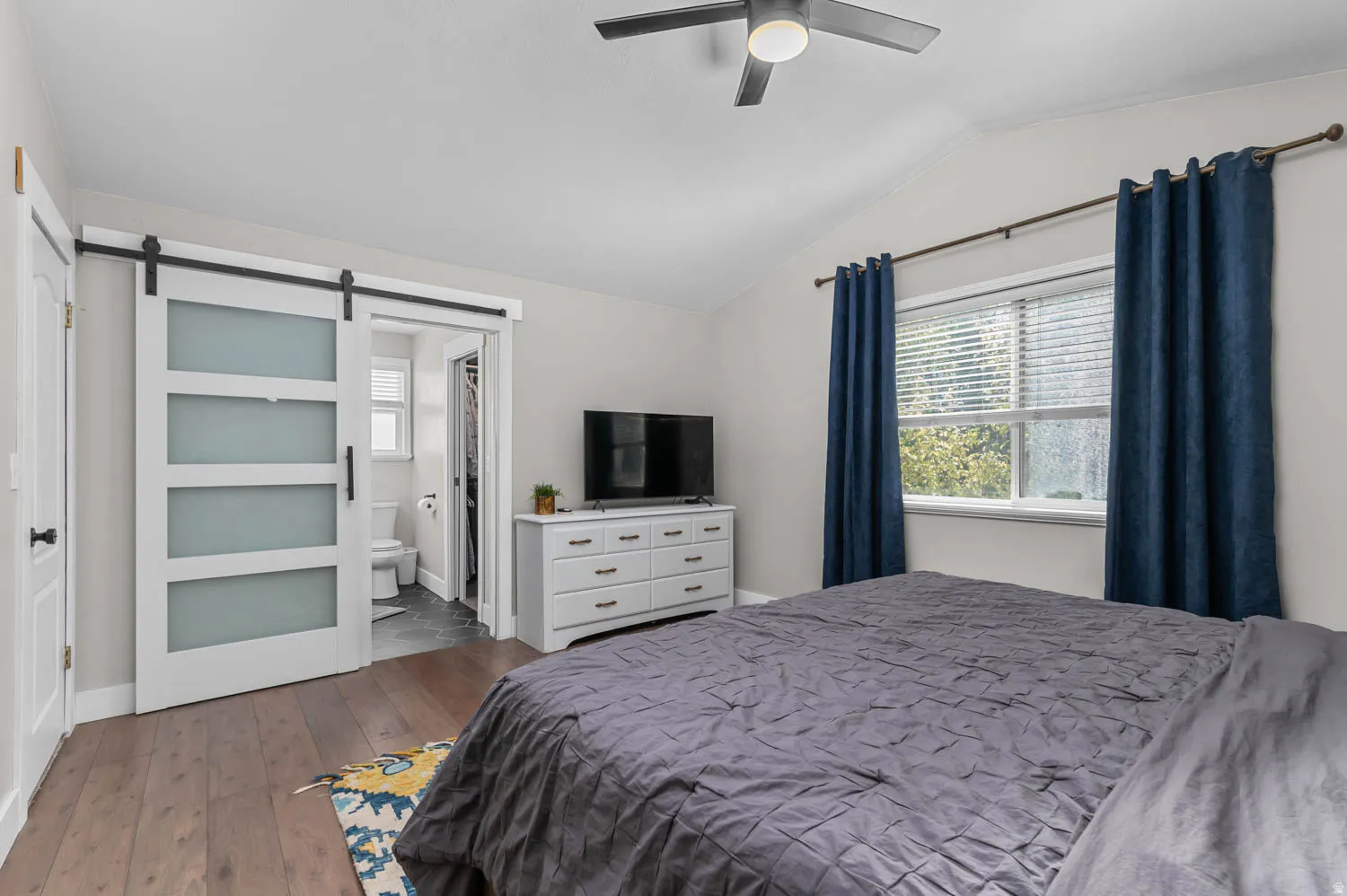 Vaulted ceiling bedroom with wood-finish flooring, a ceiling fan with integrated lighting, and a window with blinds