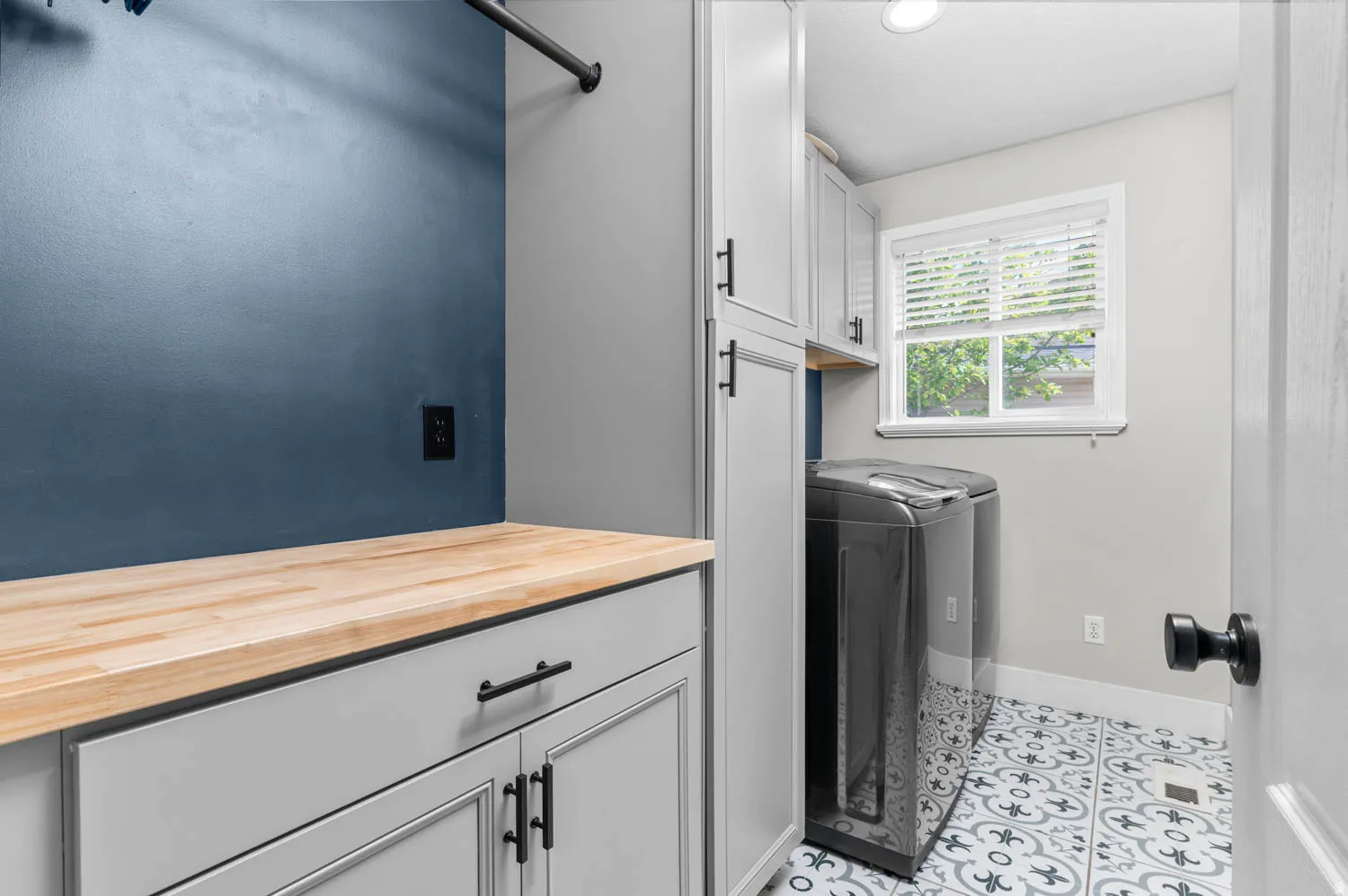 Laundry room featuring patterned floor tile, light gray cabinetry with black hardware, a wood-finish countertop, and a window with blinds