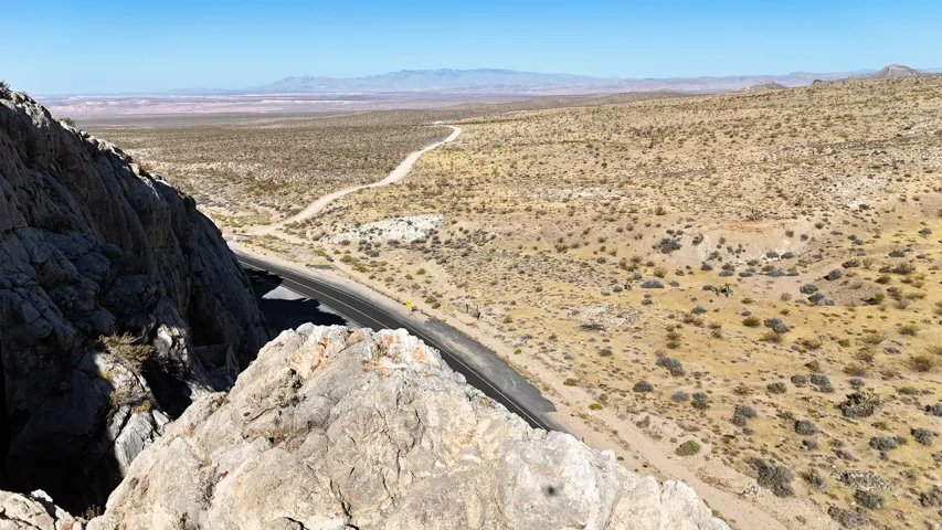 Vast desert landscape featuring a paved road winding through arid terrain, a distant dirt track, rugged rock formations, and expansive clear skies