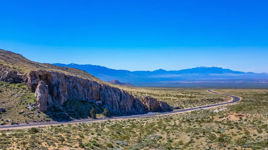 Expansive desert landscape featuring a winding paved road, rugged rock formations, and distant mountains